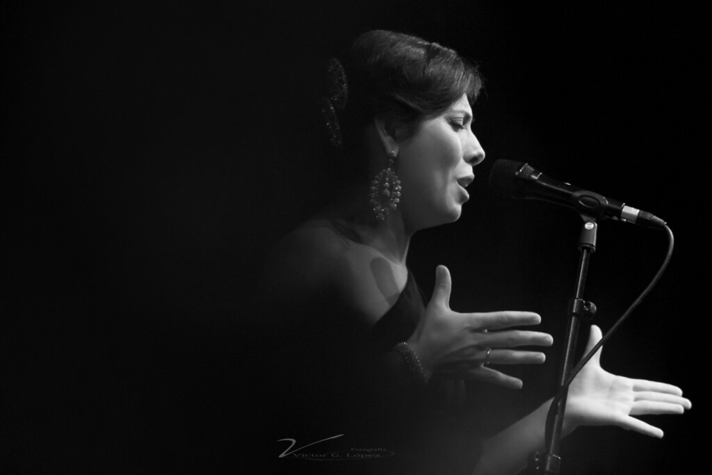 Fotografía en blanco y negro de una mujer cantando en un escenario en el concurso de flamenco de Almería. Está frente a un micrófono y parece estar interpretando con pasión, con las manos extendidas. Usa aretes grandes y lleva el cabello recogido hacia atrás.